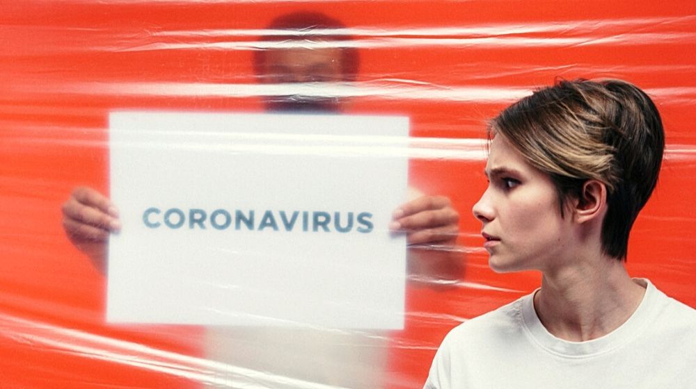 man holding paper with coronavirus behind the plastic cover and woman with worried face | WHO Clarifies That There Is “No Evidence” That Those Who Recovered from COVID-19 Are Safe from Reinfection | Featured