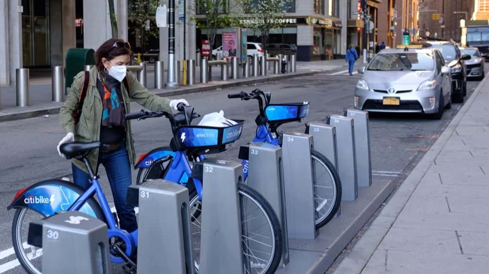 A Woman with Personal Protective Equipment Parking a Citi Bike | Robert Kaplan Says U.S. Economy’s Recovery May Depend on Success in Limiting Spread of COVID-19 | Featured