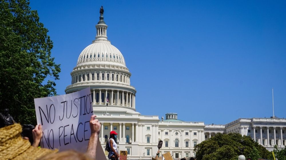 A Protestor Holds a Sign That says "No Justice No Peace" in Front of the Capitol | Grandfather of Young Boy Killed by a Stray Bullet in Washington D.C. Says “We Need the Police” | Featured