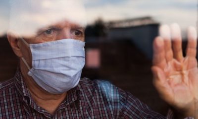 Portrait of Elderly Senior Citizen Wearing Face Mask Looking Through Room Window | COVID-19 Death Counts Up For Debate Yet Again | Featured