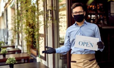 Happy waiter with protective face mask holding open sign while standing at cafe doorway-new ppp loans-ss-featured