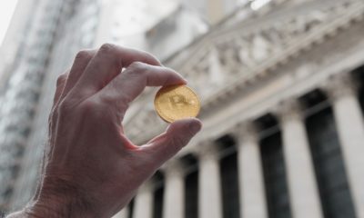 Bitcoin coin being held up outside the New York City Stock exchange building-Warming Up To Bitcoin-ss-featured