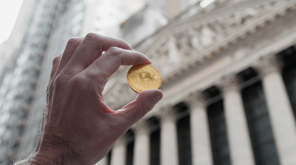 Bitcoin coin being held up outside the New York City Stock exchange building-Warming Up To Bitcoin-ss-featured