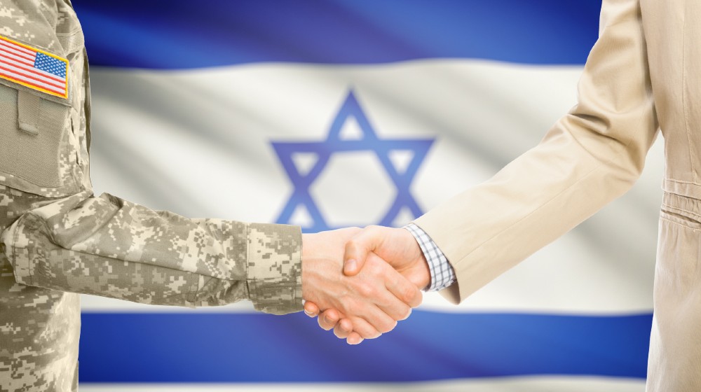 American soldier in uniform and civil man in suit shaking hands with adequate national flag on background - Israel | US Rejects UN Security Council Call For Ceasefire | Featured