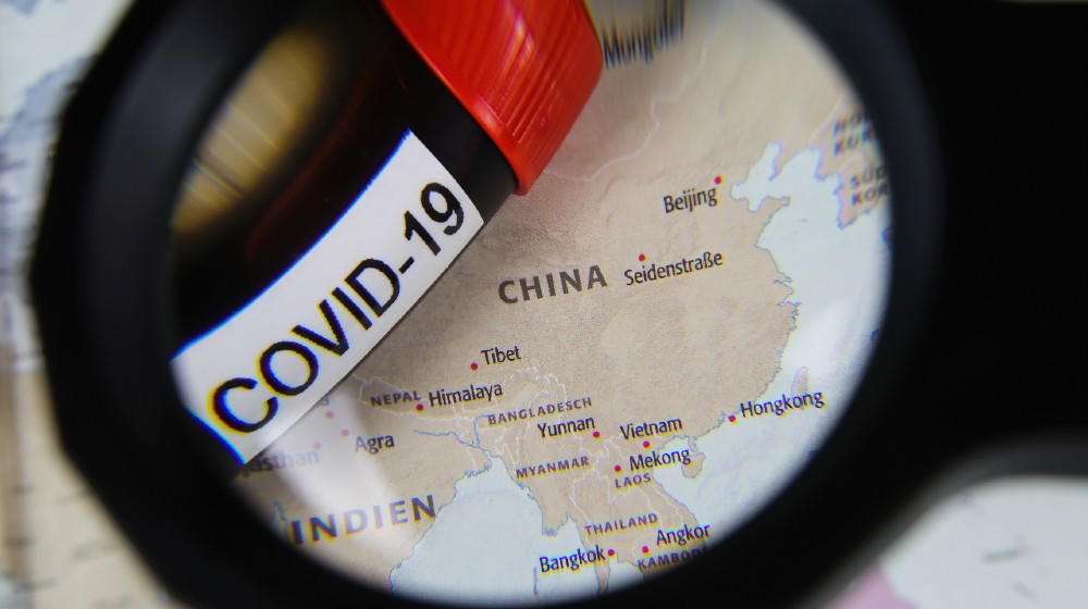 View through magnifying glass on map with focus on China and blood sample vial covid-19 | McCarthy Considers Plans to Sue China For COVID Deaths | featured