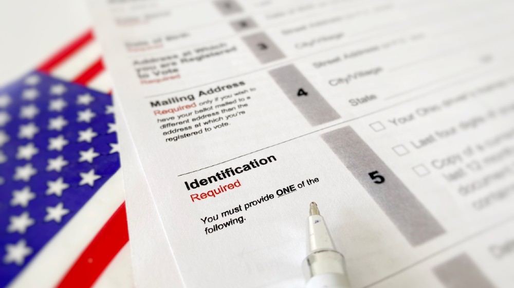 November 2019 pen pointing at identification requirements on American election application form ballot with blurry flag next to form | 78% Of Americans In Favor Of Stronger Voter Laws | featured
