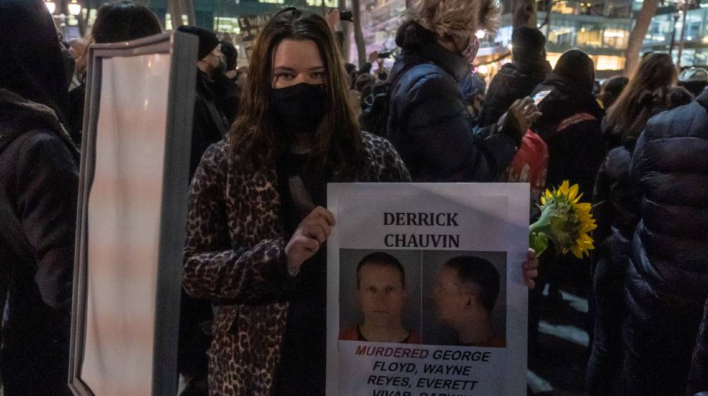 A woman holds a sign with Derrick Chauvin's picture as people gather for a George Floyd protest | Derick Chauvin: The Sacrificial Lamb | featured