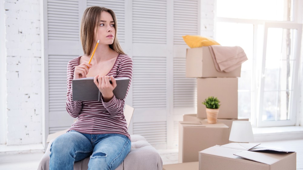 Neat and organized. Pleasant young woman sitting on the pouf in her old apartment and checking the list of items in the notebook before moving out | Important Facts About Moving Insurance | featured