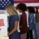 Four women of various demographics, young blonde woman in front, filling in ballots and casting votes in booths at polling station | It’s Not a Democracy if the Votes Don’t Count | featured