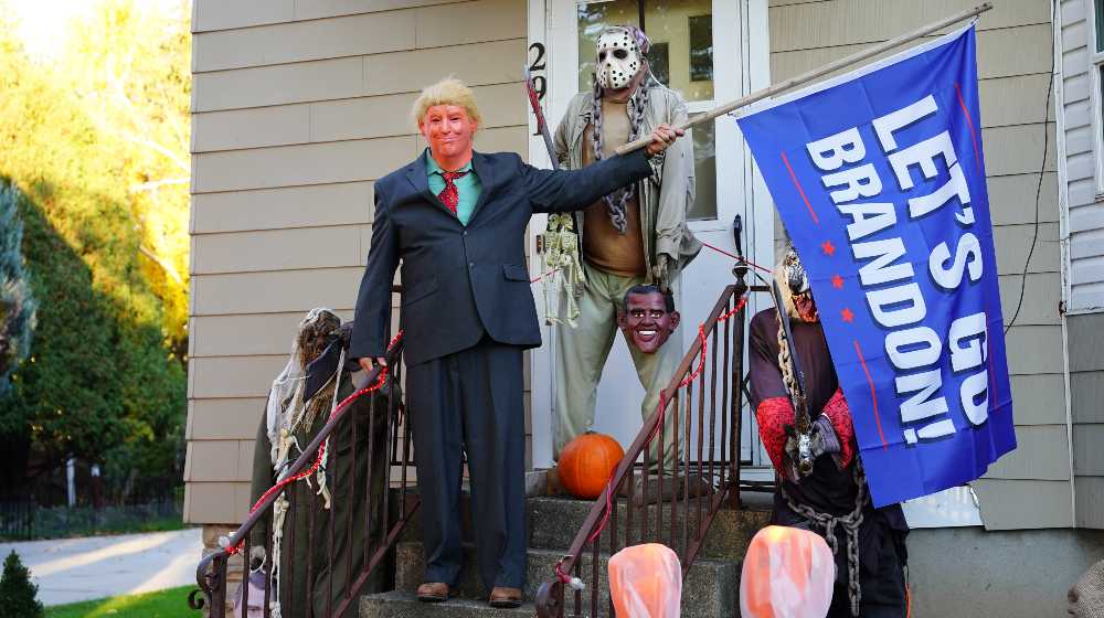 Man dressed as President Donald Trump holding a Let's Go Brandon flag for Halloween | Ron DeSantis Went to Brandon, Florida to Troll Joe Biden | featured