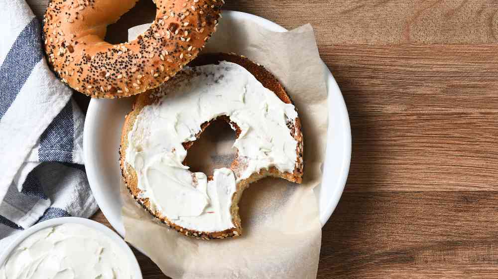 A bagel with cream cheese on a plate and a bite taken out | Kraft’s Cream Cheese Shortage Can Win Some Americans $20 | featured