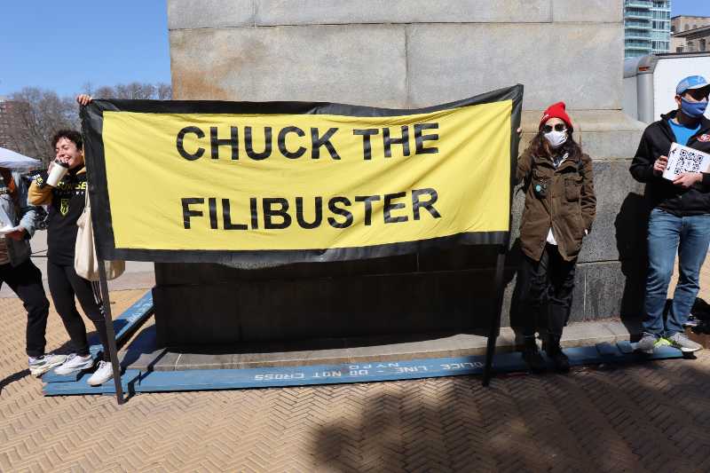Protesters wearing masks holding large bright yellow Chuck the Filibuster banner sign | Nuclear Winter