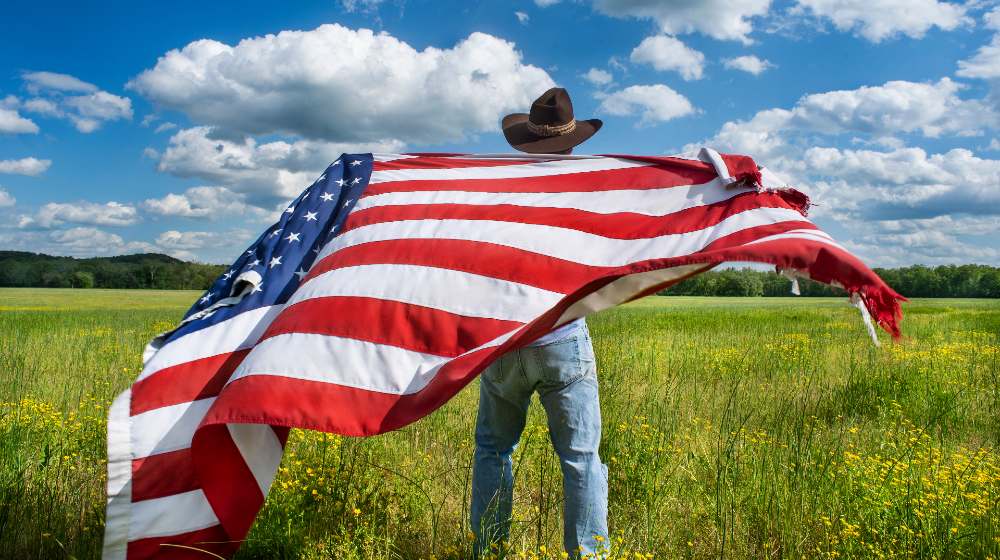 Man wearing cowboy hat waving American flag standing in grass farm-Democratic Party-SS-Featured