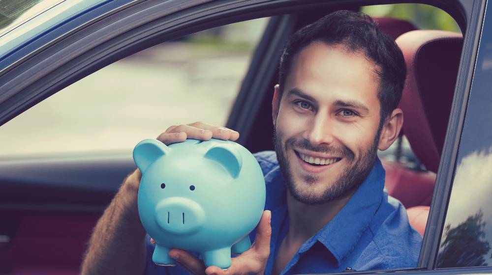 Side view happy man sitting inside his new car holding piggy bank | Democrats Want $100 Monthly Stimulus Checks For Gas | featured
