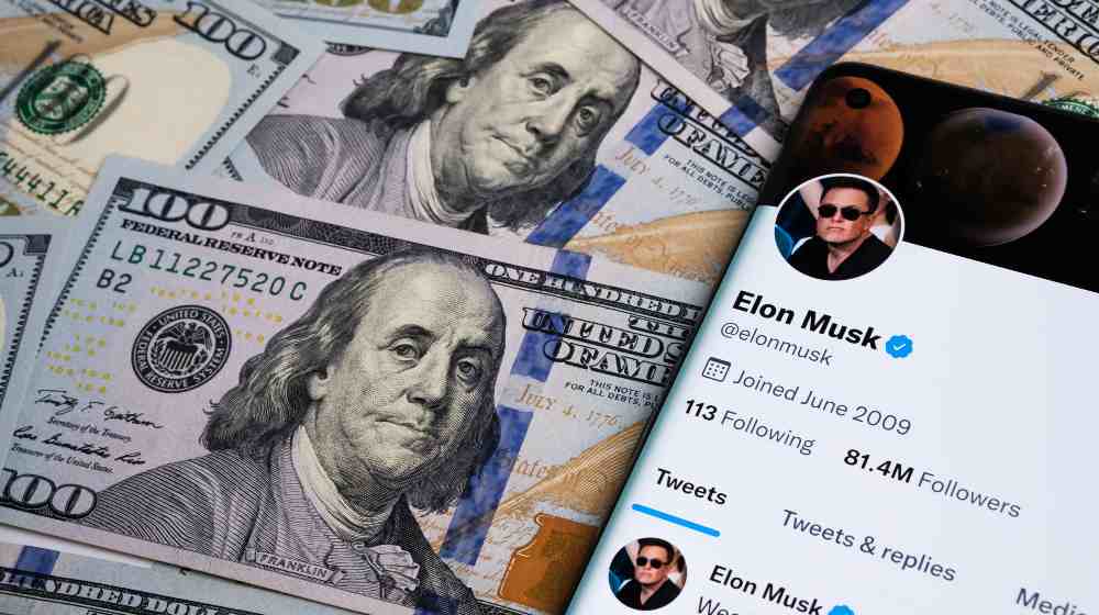 Elon Musk's Twitter account page on the smartphone which is is placed on the pile of dollar banknotes | Board Agrees to Elon Musk’s $44B Twitter Buyout Offer | featured