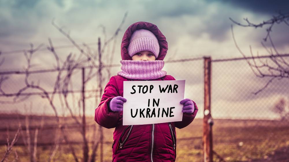 Little refugee girl with a sad look and a poster that says stop war in Ukraine | Putin is Committing Genocide in Ukraine, According to Biden | featured