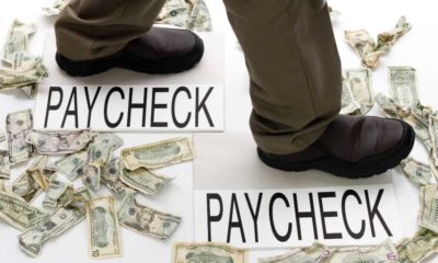Male legs walking from one paycheck to another with crumpled money scattered about on the floor | 2 out 3 American Workers Are Now Living Paycheck to Paycheck | featured