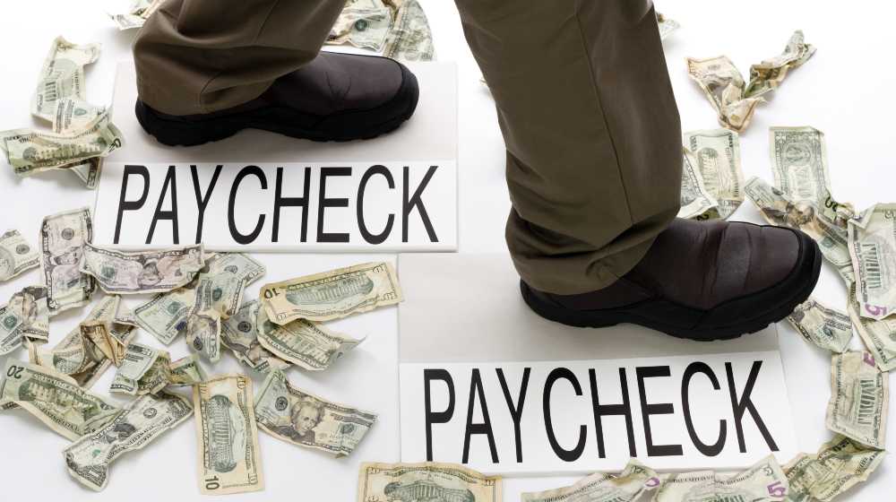 Male legs walking from one paycheck to another with crumpled money scattered about on the floor | 2 out 3 American Workers Are Now Living Paycheck to Paycheck | featured