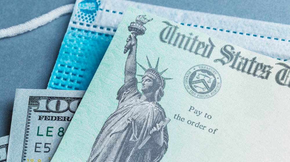 A US stimulus check on with some one-hundred-dollar bills and a medical mask-1400 Stimulus Checks-ss-featured