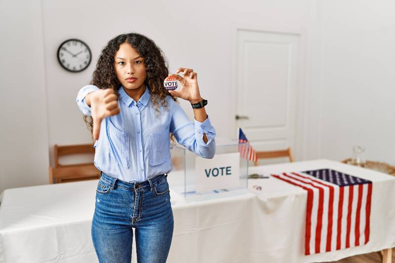 Beautiful hispanic woman standing by at political campaign by voting ballot looking unhappy-Popularity Rating