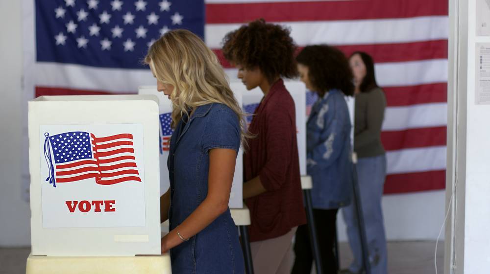 Four women of various demographics, young blonde woman in front, filling in ballots and casting votes in booths at polling station | It’s Not a Democracy if the Votes Don’t Count | featured
