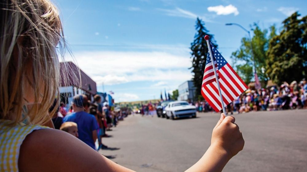 Girl Holding USA Flag at 4th of July Parade Festival Celebration | July 4 is a Time to Celebrate, Not Denigrate, the U.S.A. and its Birth | Commentary | Featured
