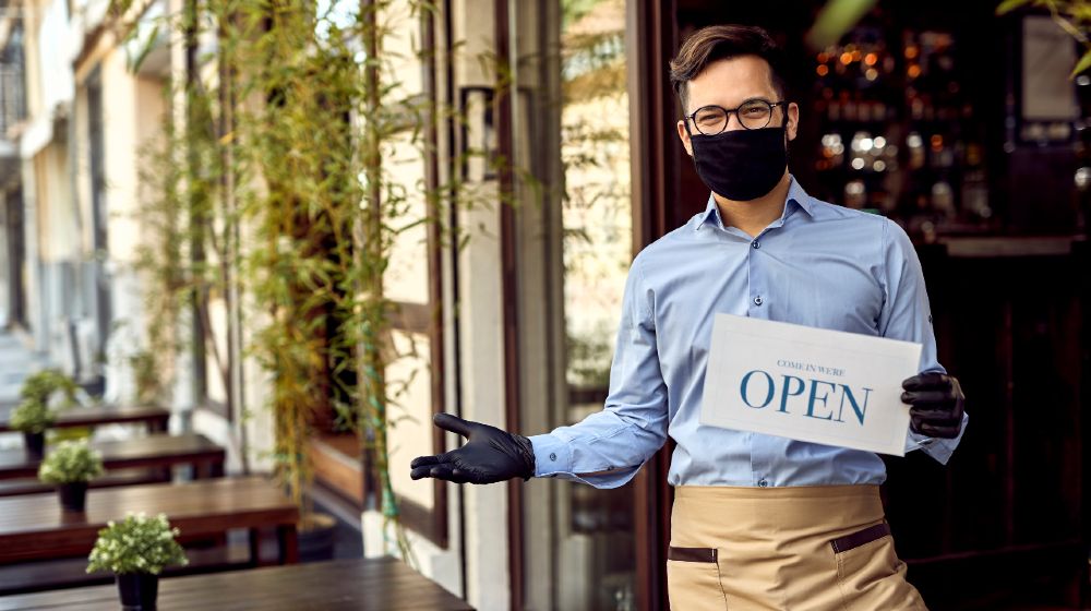 Happy waiter with protective face mask holding open sign while standing at cafe doorway-new ppp loans-ss-featured