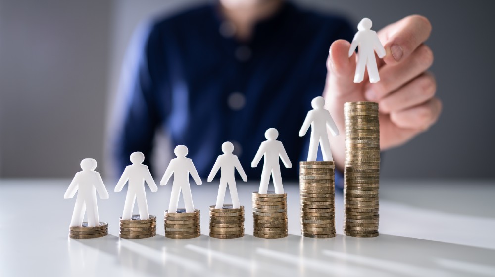 Human Hand Placing Small Human Figure On Increasing Stacked Coins Over Wooden Desk-15 Per Hour Minimum Wage-ss-featured