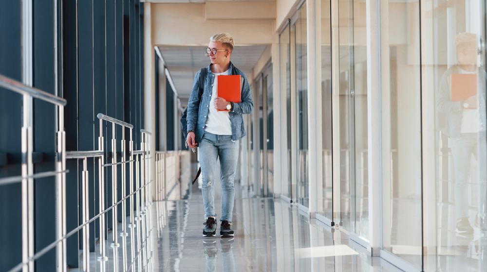 Male young student in jeans clothes is in corridor of a college with notepad in hands | Universities To Require Vaccines For Returning Students | Featured