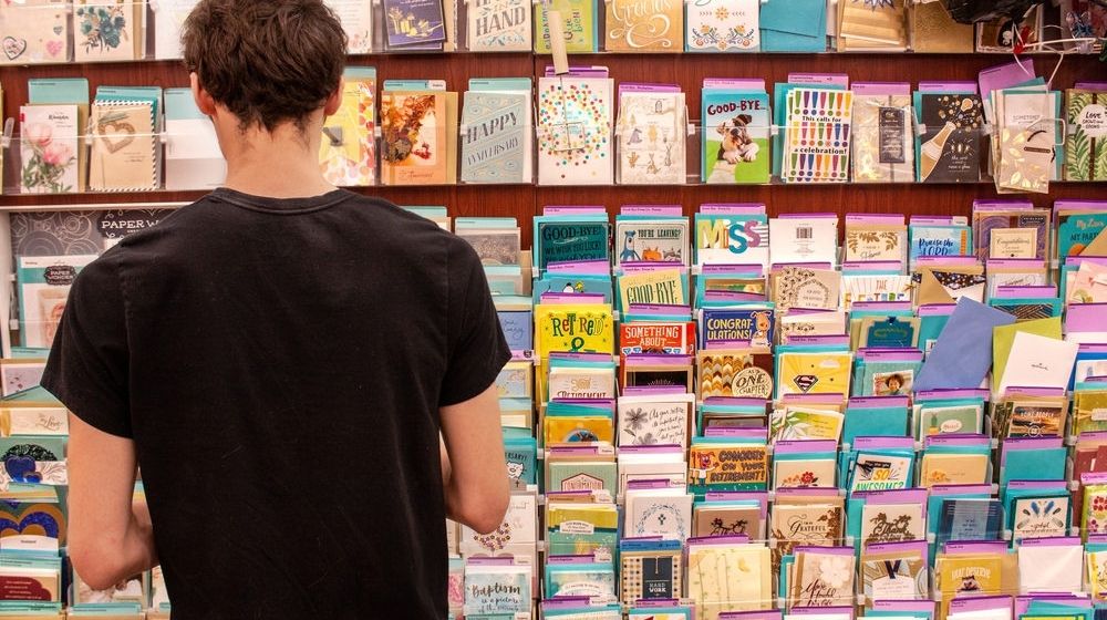 Man in Black Shirt Standing in Front of Greeting Cards Section of a Grocery Store | Greeting Card Company Hires Team of Employees Who Have Gone Through Homelessness to Design Its Cards | Featured