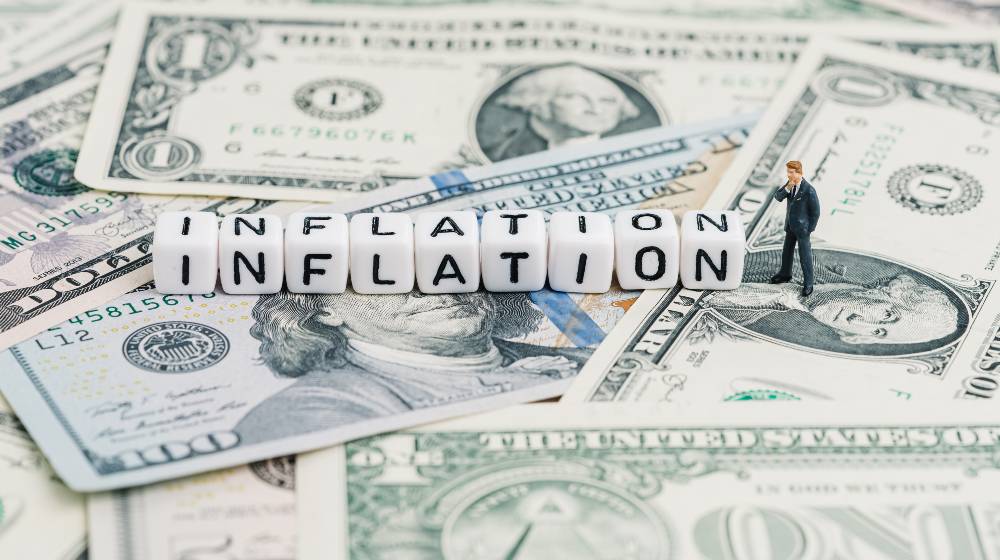 Miniature people businessman standing and wise thinking with cube block combine word Inflation on US dollar money banknote | Inflation Explained | featured