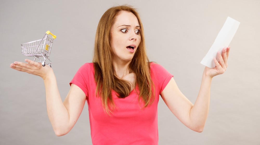 Shocked woman holding list of products or bill and small shopping cart bag trolley | Higher Grocery Prices Make Americans Buy Less, Switch Stores | featured