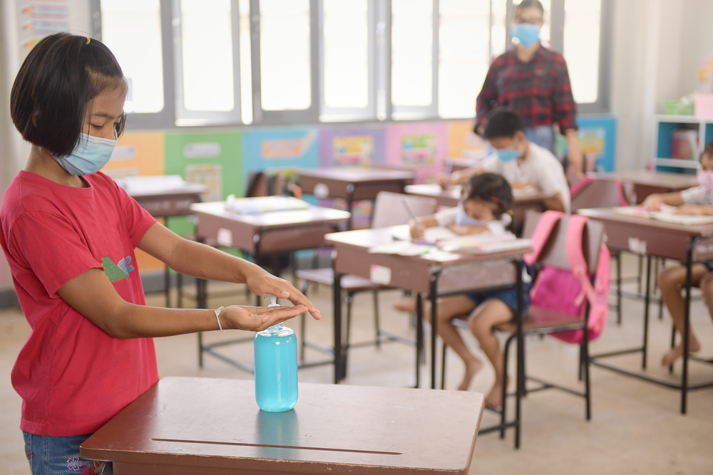 Three students and a teacher in a classroom with face mask and a girl applying hand sanitizer to her hands-Teachers’ Unions Call to Halt In-Person Learning as COVID-19 Cases Rise-ss-featured