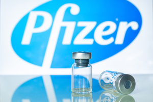 Vaccine vials in front of the Pfizer logo- Pfizer Coronavirus Vaccine Volunteer Talks About Its Side Effects-ss-Featured