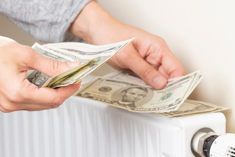 Woman's hand counting US dollars banknotes and place on heating radiator battery | Household Expenses Increased by An Average $296.45 Per Month