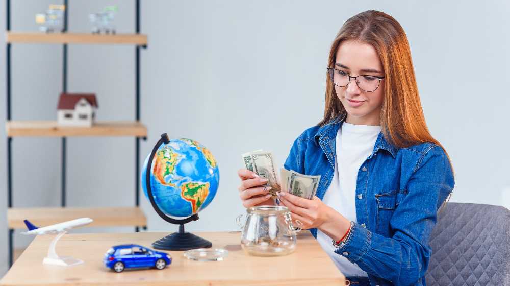Young beautiful girl counts money from a piggy bank while sitting at a table | More States Want Students To Take Personal Finance Classes | featured